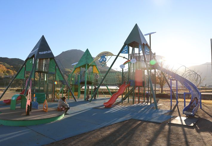 青空と山々に囲まれた公園内には大きなピラミッド型の遊具が並びます。大小の滑り台や吊り橋などで親子で遊ぶ姿が映った写真です。